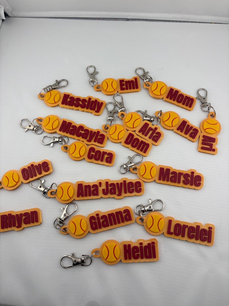Pu&ograve; includere: Una collezione di portachiavi personalizzati gialli e marroni, ognuno con un'icona di palla da softball e un nome o titolo tra cui Kassidy, Emi, Mom, MaCayla, Aria, Dom, Ava, Ari, Olive, Cora, Ana'Jaylee, Marsie, Rhyan, Gianna, Lorelei e Heidi.