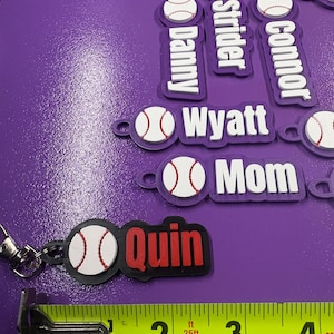 Pu&ograve; includere: Una collezione di portachiavi personalizzati con una pallina da baseball bianca con cuciture rosse attaccata a un'etichetta rettangolare. Le etichette riportano vari nomi come Quin, Mom, Wyatt, Connor, Strider e Danny. Un metro a nastro giallo indica una lunghezza di circa 8,9 cm.