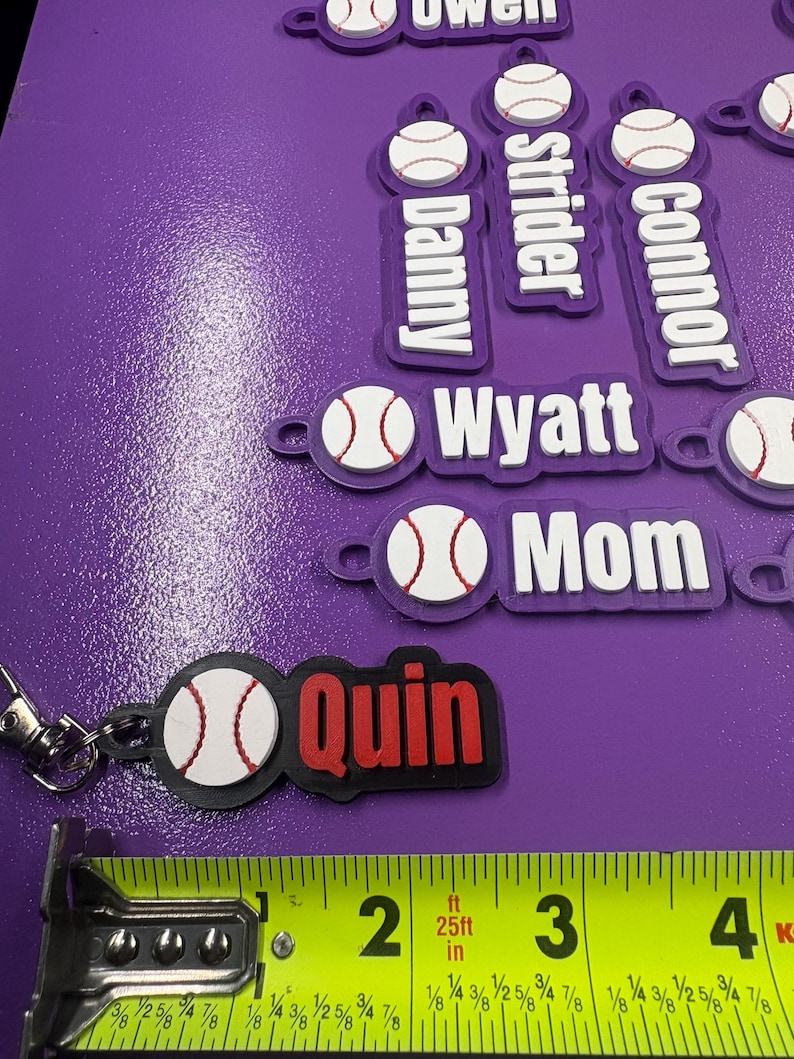 Pu&ograve; includere: Una collezione di portachiavi personalizzati con una pallina da baseball bianca con cuciture rosse attaccata a un'etichetta rettangolare. Le etichette riportano vari nomi come Quin, Mom, Wyatt, Connor, Strider e Danny. Un metro a nastro giallo indica una lunghezza di circa 8,9 cm.