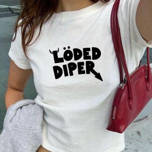 May include: White t-shirt with the text "LÖDED DIPER" in black, paired with gray trousers and a pink belt. A red handbag hangs from the shoulder. A gray sweater is held in the arm.