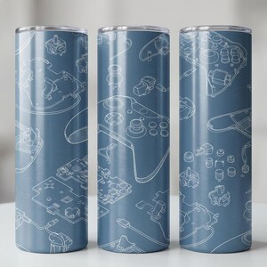 May include: Three blue stainless steel tumblers featuring a white schematic design of video game controllers and components. The design includes controllers, circuit boards, and buttons. The tumblers are cylindrical with a silver rim.