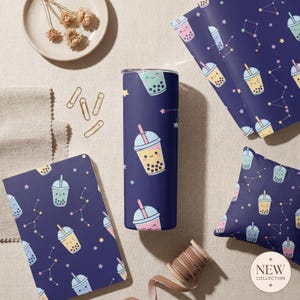 Può includere: Un bicchiere, un taccuino, carta da regalo e un cuscino blu navy con un motivo ripetuto di bevande al tè alle bolle e costellazioni. Il bicchiere è cilindrico e presenta un design a tè alle bolle. Altri oggetti includono un piatto con fiori secchi e graffette.
