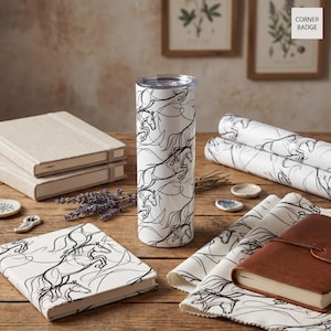 May include: A white tumbler with a black horse design stands on a wooden table, surrounded by matching notebooks, fabric, and a journal. The tumbler is cylindrical and has a clear lid. The horse design is a continuous line drawing.