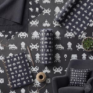 Può includere: Una collezione di oggetti con un motivo pixelato di space invaders bianchi su sfondo grigio scuro. Include un bicchiere, un taccuino, carta da regalo, un campione di tessuto, un cuscino e una pianta succulenta.