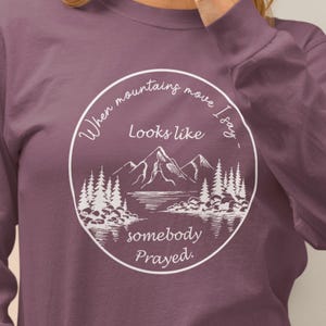 Puede incluir: Una camiseta de manga larga de color morado con un diseño gráfico blanco que dice "Cuando las montañas se mueven, digo, parece que alguien ha rezado."