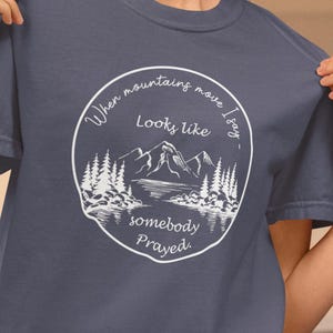 May include: A navy blue t-shirt with a white graphic of a mountain range and a lake. The graphic is surrounded by a circle with the text "When mountains move I say, Looks like somebody prayed."