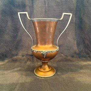 May include: A copper trophy with silver handles and a decorative base. The trophy has an inscription that reads "First Annual Bowling Tournament".