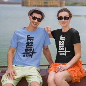 Puede incluir: Dos personas con camisetas a juego con el texto "Jes&uacute;s es Rey" y un gr&aacute;fico de cruz. El hombre lleva una camiseta azul claro y la mujer una camiseta negra. Ambos sonr&iacute;en y miran a la c&aacute;mara.