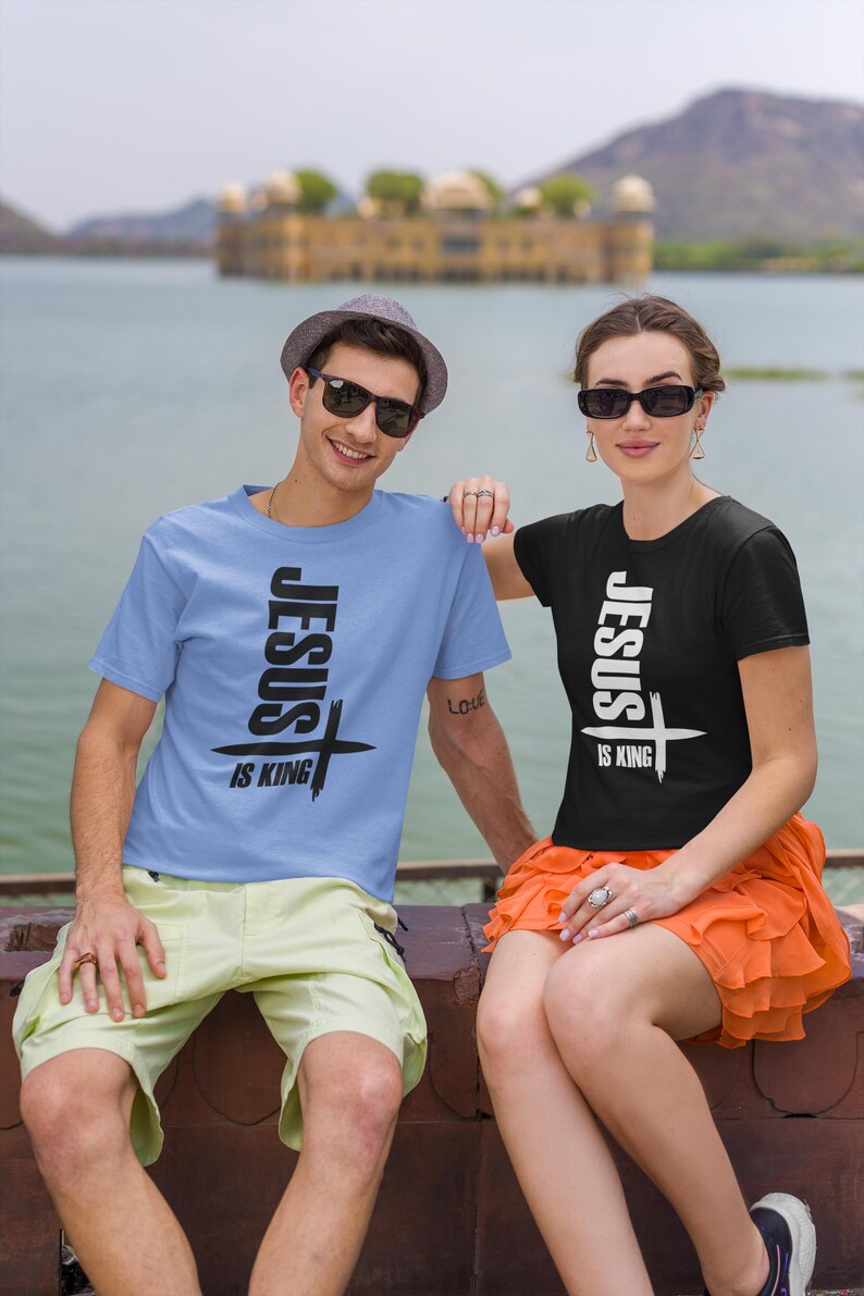 Puede incluir: Dos personas con camisetas a juego con el texto "Jes&uacute;s es Rey" y un gr&aacute;fico de cruz. El hombre lleva una camiseta azul claro y la mujer una camiseta negra. Ambos sonr&iacute;en y miran a la c&aacute;mara.