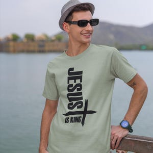 Puede incluir: Una camiseta verde claro con un gr&aacute;fico negro que dice "JESUS IS KING" con una cruz.