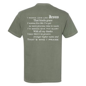 Puede incluir: Camiseta verde oliva con texto blanco en la espalda. El texto incluye la palabra "Jesus" y un verso religioso. La camiseta tiene mangas cortas y cuello redondo.
