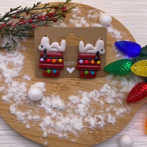 May include: A pair of Snoopy-themed earrings. Each earring features a white Snoopy figure lying on a red doghouse decorated with colorful Christmas lights. The earrings are displayed on a brown card, with a wooden background and decorative lights.