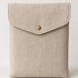 May include: A rectangular sleeve case made of light beige textured fabric. The case features a flap closure secured with a single centred brass-coloured metal snap button. The edges are finished with visible stitching along the perimeter of the flap.