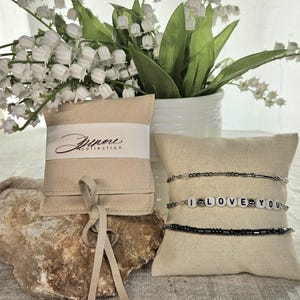 May include: An image showcases three beaded bracelets on a beige pillow, one spelling "I LOVE YOU." A beige pouch with the text "Zimore collection" is also present. The background includes a white vase with white flowers and green leaves.