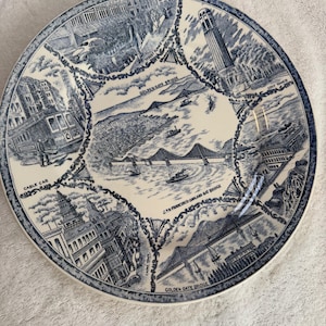 May include: A round ceramic plate featuring blue and white illustrations of San Francisco landmarks. The center depicts the Golden Gate Bridge and San Francisco-Oakland Bay Bridge. Surrounding panels show a cable car, Coit Tower, and Chinatown.