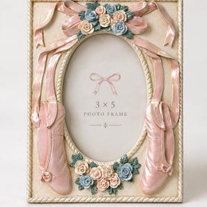 May include: A rectangular photo frame with an oval center, featuring a cream-colored border with a rope-like edge. The frame is decorated with sculpted pink ballet slippers, pink ribbons, and clusters of small pink and blue roses. Text reads 3 x 5 PHOTO FRAME.