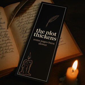 以下が含まれることがあります： 「the plot thickens some pages burn slower」のテキストと羽のイラストが描かれた黒いブックマーク。背景には、火の灯ったろうそくと、手書きのテキストが書かれた開いた本があります。ブックマークは長方形です。
