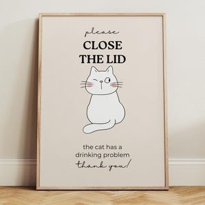 May include: A framed print with a light beige background features a cartoon cat winking. Above the cat, the text reads "please CLOSE THE LID." Below the cat, the text says "the cat has a drinking problem thank you!"