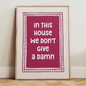 May include: A rectangular wall art print in a light wood frame featuring a deep magenta background with a white hand-drawn border. The text in white reads: in THIS HOUSE WE DON'T GIVE A DAMN. The design is centered on a white paper background.