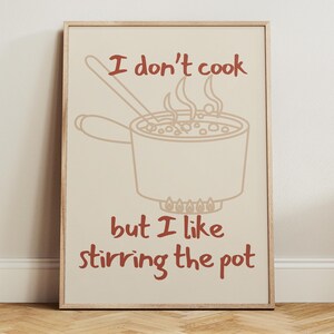 May include: A framed art print with a beige background and a brown outline illustration of a pot on a stove. The text reads, "I don't cook but I like stirring the pot."