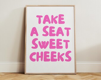 May include: A framed art print with the phrase "TAKE A SEAT SWEET CHEEKS" in bright pink, bold, rounded letters on a white background. The frame is a light wood color. The print is displayed against a neutral wall and a wooden floor.
