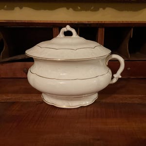 May include: A white porcelain chamber pot with a lid and handle. The pot has gold trim and a decorative design around the rim. The pot is sitting on a dark wooden surface.