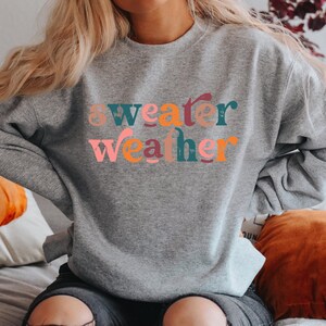 May include: A heather gray crewneck sweatshirt featuring the text "sweater weather" printed across the chest. The letters are rendered in a distressed, multi-colored font with shades of pink, teal, orange, and dark red throughout the design.