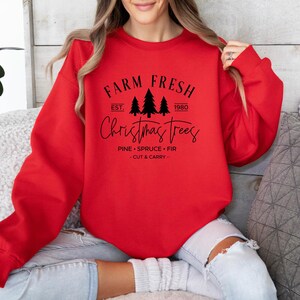 May include: A bright red crewneck sweatshirt featuring black text and graphics. The design includes the words FARM FRESH, EST. 1980, and Christmas Trees, along with three pine tree silhouettes and the text PINE, SPRUCE, FIR, CUT & CARRY.