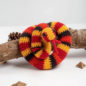 May include: A crocheted snake toy in a coiled position. The snake features a vibrant color scheme of red, yellow, and black stripes. It has large black eyes and a red tongue. The toy is placed on a white surface with a piece of wood and a pinecone.