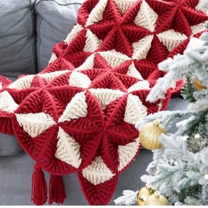 May include: A red and cream-colored crocheted blanket with a starburst pattern. The blanket is draped over a gray sofa. The blanket has tassels at the bottom. A Christmas tree with gold ornaments is in the background.