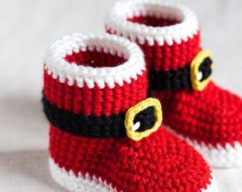 May include: A pair of handmade crochet baby booties in a Santa Claus theme. The booties are primarily red with white trim around the top and soles. Black belts with yellow buckles are wrapped around the ankles.