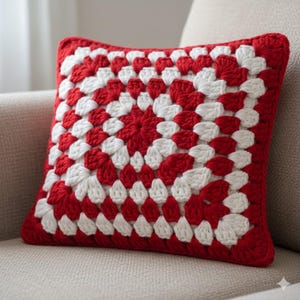 May include: Square crochet pillow in red and white. The pillow features a granny square design with a red border. The pattern is a repeating sequence of red and white squares, creating a textured surface. The pillow is placed on a neutral-colored sofa.