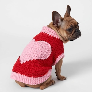 Peut inclure: Un pull pour chien rouge et rose au crochet, orné d'un grand cœur rose sur le dos. Le pull a un col et un ourlet roses côtelés. Le chien est un bouledogue français marron, assis et regardant sur le côté.