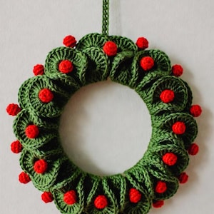 May include: A handmade green crochet wreath with red berry accents. The wreath is circular, with a loop for hanging. The red berries are evenly spaced around the wreath, adding a festive touch. Ideal for holiday decor.