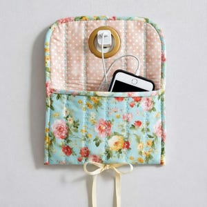 May include: A floral-patterned wall-mounted phone charging station. The top section has a pink polka-dot fabric, while the pocket features a blue floral design. A white phone is in the pocket, with a charger plugged into a gold outlet. A cream ribbon bow hangs below.