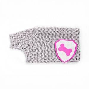 Puede incluir: Un suéter para perros de punto gris claro. Tiene un parche rosa y blanco en forma de escudo con un diseño de hueso rosa. El suéter está diseñado para adaptarse al cuerpo de un perro, con aberturas para las patas y el cuello.