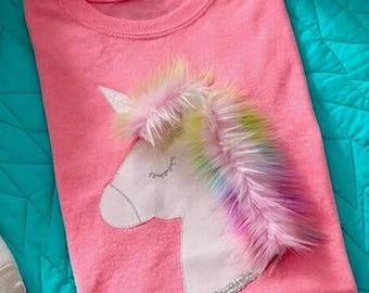 May include: A pink t-shirt featuring a white unicorn design with a silver glitter outline and a colorful faux fur mane. The shirt is folded on a turquoise quilted surface.