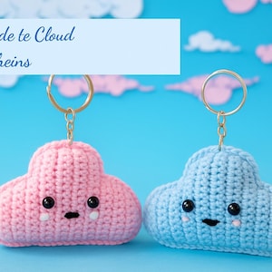 May include: Two handmade cloud-shaped keychains, one pink and one blue, with black eyes and smiling faces. The keychains are attached to gold-colored rings. The text "Claude te Cloud keychains" is in a light blue rectangle above the keychains.
