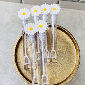 May include: A collection of clear bubble wands, each topped with a white daisy with a yellow center. The wands are arranged on a gold-colored tray. The wands are clear with white handles.