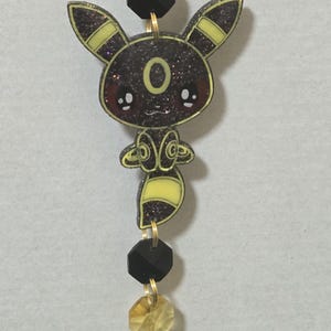 May include: A hanging ornament featuring a glittery black and yellow creature charm with pointed ears and a tail, connected by gold-tone metal rings to faceted black and translucent yellow beads, ending in a large, teardrop-shaped yellow crystal.