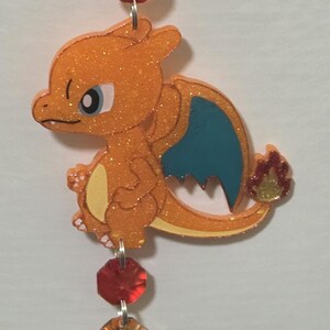 Può includere: Un ornamento sospeso raffigurante un personaggio drago arancione glitterato con ali blu e una fiamma sulla coda, sospeso da una catena verticale di perline sfaccettate gialle, arancioni e rosse, che termina con un grande cristallo rosso scuro a forma di goccia.