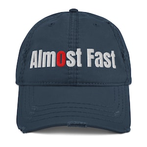 May include: A navy blue distressed baseball cap featuring the embroidered white text Almost Fast with the letter o highlighted in red. The cap has a six-panel construction with a curved brim and visible frayed edges along the seams and visor.
