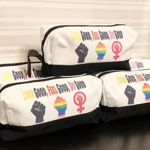 May include: A stack of white and black zippered cosmetic bags. Each bag has the text "LOOK GOOD, FEEL GOOD, DO GOOD" in colorful letters, along with a black fist, a rainbow hand, and a female symbol.