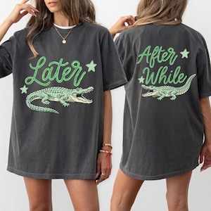 May include: Dark grey t-shirts featuring a green alligator design and the phrases "Later" and "After While". The front of the shirt displays "Later" with an alligator and stars. The back of the shirt shows "After While" with an alligator and stars.