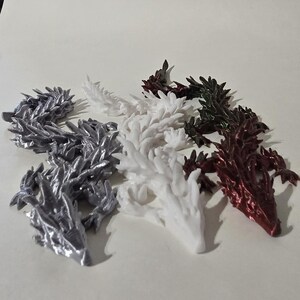 May include: An assortment of dragon figurines in various colors, including silver, white, dark green, and burgundy. The dragons are intricately detailed and appear to be 3D-printed, showcasing a fantasy theme.