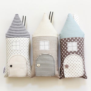 May include: Three plush toy houses in the shape of pillows. The houses are made of different fabrics with various patterns. The first house is white with black stripes, the second is beige with brown stripes, and the third is brown with white stars and blue stripes.