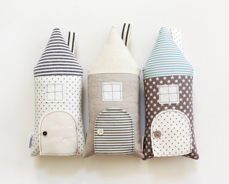 May include: Three plush toy houses in the shape of pillows. The houses are made of different fabrics with various patterns. The first house is white with black stripes, the second is beige with brown stripes, and the third is brown with white stars and blue stripes.