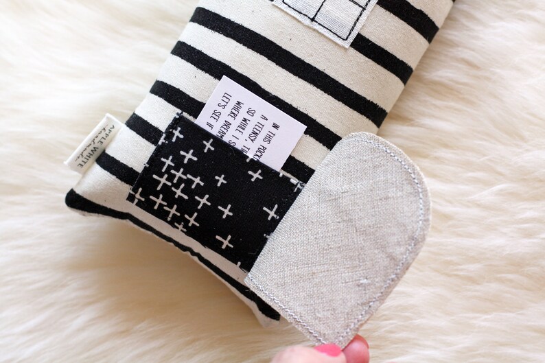 May include: A black and white striped pillow with a small pocket on the side. The pocket has a white fabric with a black cross pattern. A white tag is sticking out of the pocket with the text "IN THIS POCK A TEENSY, TH SO WHILE I S WHERE DREAM LET'S SEE IF". The pillow has a heart-shaped flap on the side.