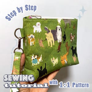 Puede incluir: Un estuche de tela verde con cremallera y un patrón repetitivo de varias razas de perros en múltiples colores. El estuche incluye una correa de muñeca a juego sujeta con un mosquetón de metal plateado y una cremallera blanca.