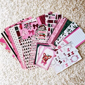 May include: A selection of pink and white Christmas paper ephemera. The collection includes patterned papers, gift tags with a deer illustration, stickers, and notepads. Some items feature bows, Christmas trees, and the text "Merry Christmas."